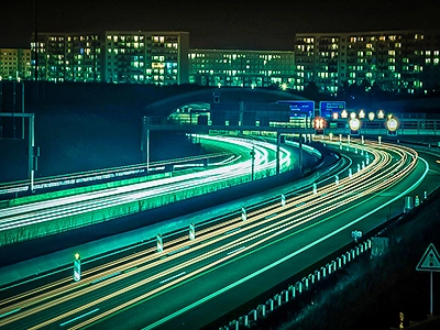 Ein beeindruckendes Farbfoto einer Autobahn bei Nacht in Jena, aufgenommen mit Langzeitbelichtung. Die Scheinwerfer und Rücklichter von fahrenden Autos erzeugen leuchtende, farbige Streifen (grünlich-weiße und gelb-orangefarbene Lichtbahnen) entlang der Fahrbahnen. Im Hintergrund sind Gebäude mit erleuchteten Fenstern unter einem dunklen Himmel zu sehen.
